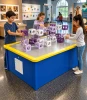 LiteZilla AeroCube Constructor Table - Blue, children build structures with purple and white modular cubes on a blue interactive construction table with a bright yellow rim in a museum.