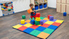 Children's Factory Patchwork Activity Mat and Foam Blocks Set, Rainbow, with a young girl playing on colorful foam blocks on a vibrant patchwork mat in a preschool classroom.