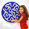 PlayScapes SpinMazera Activity Wall Toy - Choose Color, a young girl smiling and touching a circular maze game with blue and white pathways mounted on a wall.