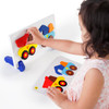 Guidecraft Construction Truck Sort and Match Game, a child's hand placing colorful wooden truck pieces onto a white matching board.