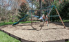 Frame It All Classic Sienna Curved Playground Border Border 16' - 1" Profile, a sturdy and durable brown landscape edging border surrounding a children's playground area with a slide and swings in a backyard setting.
