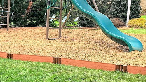 Frame It All Classic Sienna Straight Playground Border Kit 32', 2-inch profile, installed around a playground area with wood chips and a green slide, creating a safe and defined boundary.
