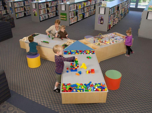 Playscapes 3-Branch Pinwheel Triple Discovery Table, a wooden activity table with three distinct sections filled with colorful building blocks, bead mazes, and train sets, used by children for educational play in a library setting.
