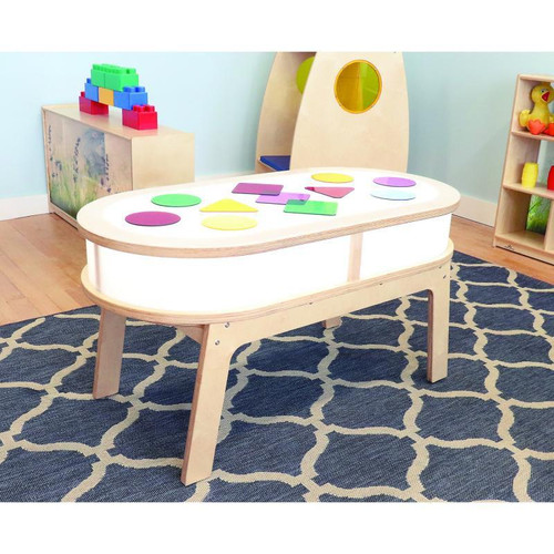 Whitney Brothers Whitney Plus Radiant LED Light Table, a children's activity table with a glowing white surface, colorful geometric shapes on top, and wooden legs, in a playroom setting with toys and a patterned rug.