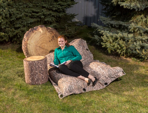 Playscapes Woodsmen Naturescape Lounger, outdoor log-inspired lounge seat on grass surrounded by pine trees with a woman relaxing and reading a book.