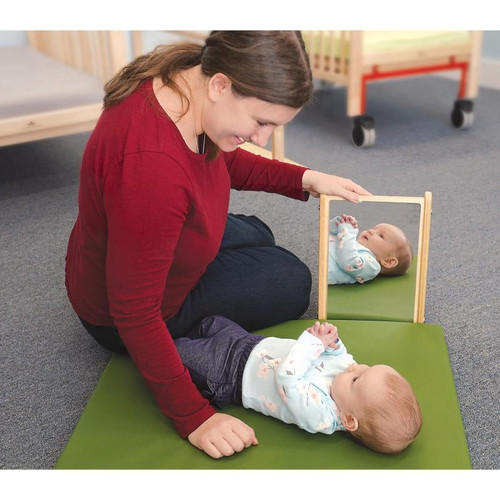 Whitney Brothers Infant Mirror Stand, a wooden baby mirror stand placed on a green mat with an infant lying on the mat and a caregiver sitting beside, engaging with the baby through the mirror.
