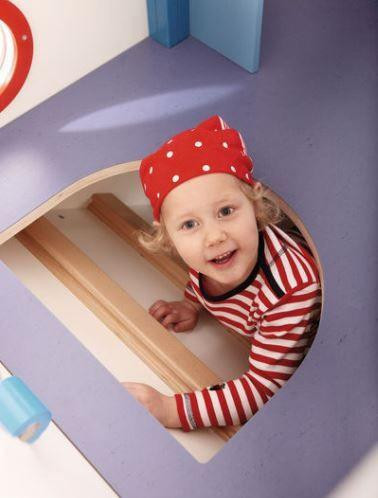 HABA Pro Voyaging Pirate Ship Playhouse, a little girl smiling and peeking through a porthole in the playhouse.