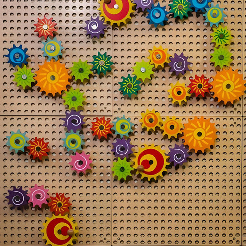 LiteZilla Kidworx Gizmo Wall Gears Wall Activity, colorful interlocking gears mounted on a beige perforated wall.