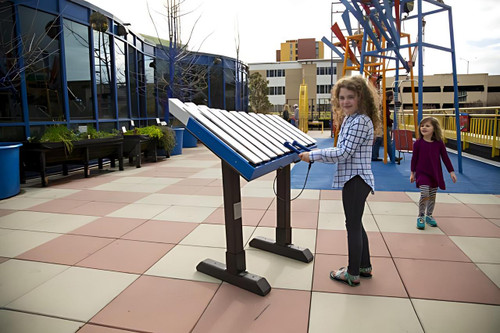 Freenotes Yantzee Outdoor Metallophone, a large outdoor metallophone instrument with metal bars, being played by a girl on a playground.