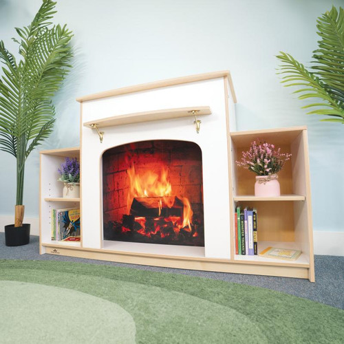 Whitney Brothers Warm and Welcoming Fireplace - White, a children's fireplace with a cozy fire display, white finish, wooden shelves with books and pink flower pot, placed in a room with green plants and a green rug.