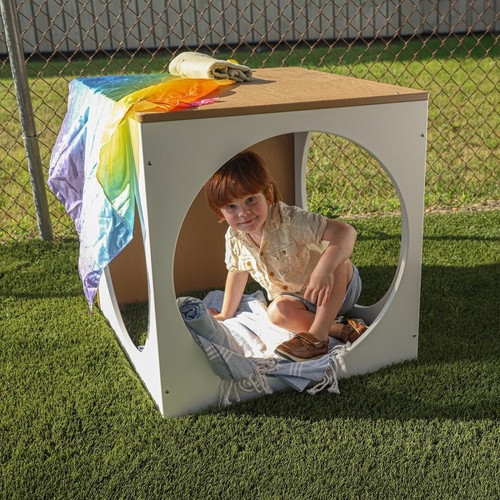 Whitney Brothers outdoorED™ Play House Cube, a children's playhouse with circular openings, featuring a child sitting inside, outdoors on grass with a rainbow cloth draped over the top.