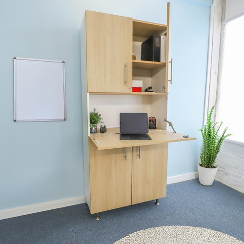 Whitney Brothers elevatED™ Workstation Cabinet, light wood finish with adjustable upper shelves and a fold-down desk area, situated in a modern office space with blue walls and a potted plant.
