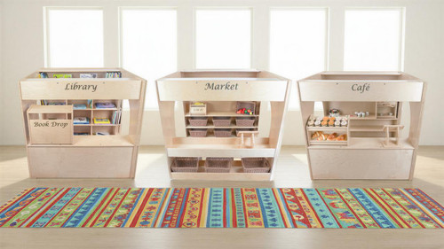 Wood Designs Town Collection - Library, Cafe and Market Playhouse Set, wooden playhouse set with labeled sections for library, market, and cafe, featuring shelves, baskets, and pretend food items, placed on a colorful patterned rug in a bright room.