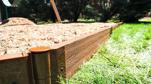 Frame It All Classic Sienna Straight Playground Border Kit 32' - 1" Profile, close-up view of the brown wood-like plastic border edge installed on grass, with mulch inside the bordered area and trees in the background.