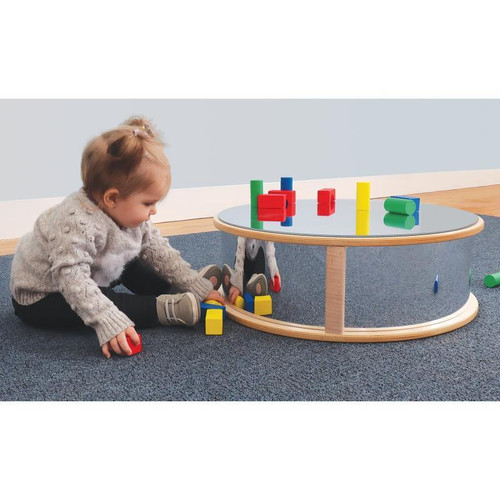 Whitney Brothers Round Infant Floor Mirror, an oval-shaped wooden mirror with a reflective surface, placed on a carpeted floor in a playroom with a young child playing with colorful building blocks nearby.