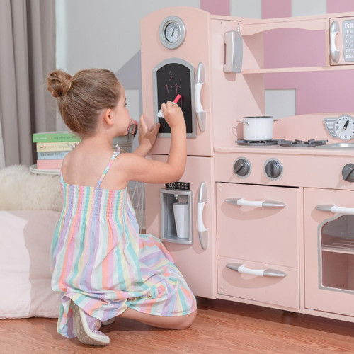 Teamson Kids Little Chef Westchester Retro Pink Play Kitchen, a pink wooden play kitchen with stove, oven, and tiny kitchen appliances, being used by a young girl drawing on a chalkboard.