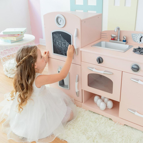 Teamson Kids Little Chef Fairfield Retro Pink Play Kitchen, a pink toy play kitchen with sink, stove, oven, and chalkboard, featuring a young girl writing on the chalkboard in a playroom setting.