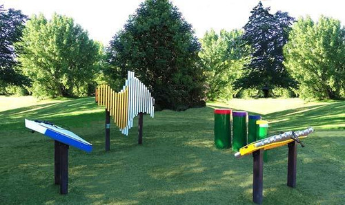 Freenotes Deluxe Ensemble Musical Playground Set, outdoor musical instruments including xylophones, drums, and metallophones arranged in a scenic park setting with green trees and grass.