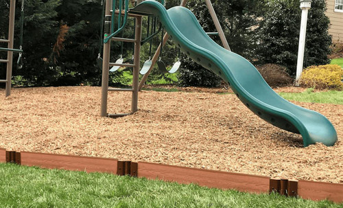 Frame It All Classic Sienna Straight Playground Border 16' - 1" Profile, a beige landscape timber with a curved top, installed around a children's playground area with a slide and a swing set in the background.
