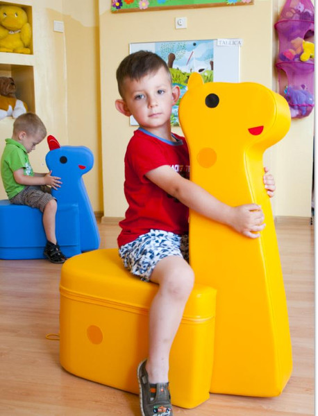 Moje Bambino Giraffe Foam Seat Pouf, yellow foam giraffe-shaped children's seat with a boy sitting on it and hugging it in a playroom setting.