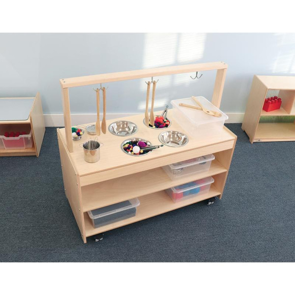 Whitney Brothers Mobile Sensory Play Kitchen, wooden sensory play kitchen with stainless steel bowls, wooden utensils, and sensory toys on a light wooden table with storage shelves.