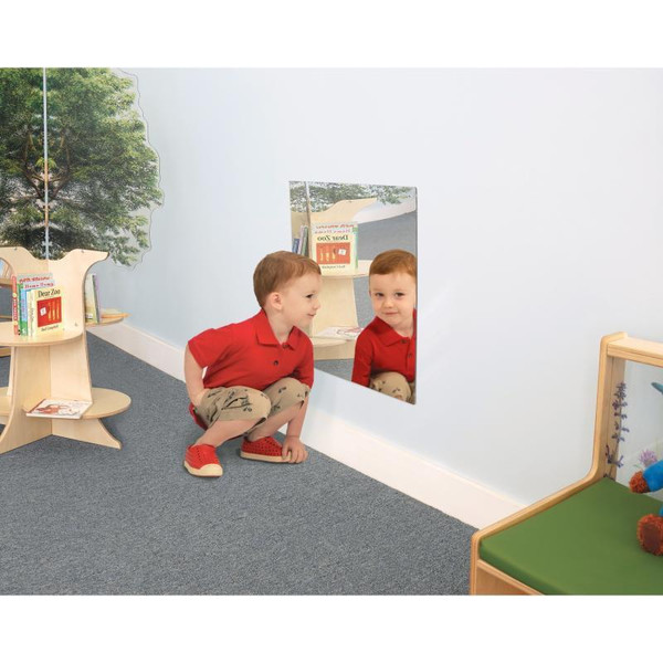 Whitney Brothers Shatterproof Acrylic Square Mirror, a child squatting in front of the mirror reflecting his face, in a playroom setting with bookshelves and a green cushioned bench.