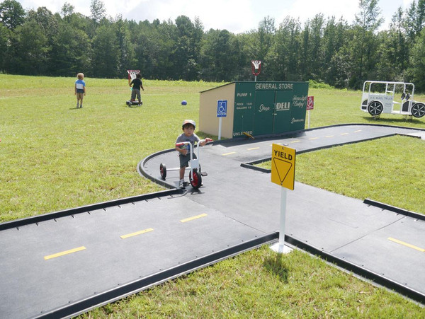 Infinity Playgrounds Atlanta Crazy Eight Trike Track, children riding tricycles and playing on an outdoor mini road with ramps, traffic signs, and mock store structures.