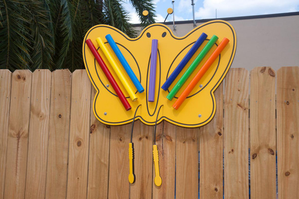 Playmore Designs Butterfly Music Chimes, colorful butterfly-shaped outdoor musical instrument with plastic chimes in red, yellow, blue, purple, green, and orange, mounted on a wooden fence.