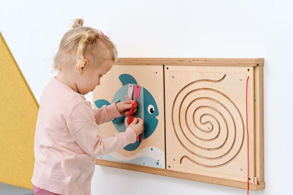 Moje Bambino Frame For Fine Motor Skills Wall Panels, a child is engaging in fine motor skill activities with a colorful fish-themed wooden wall panel.