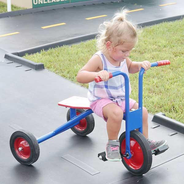 Infinity Playgrounds Infinity Toddler Tricycle, young girl riding and steering the blue and red tricycle on a paved surface outdoors.