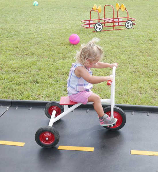 Infinity Playgrounds Infinity Toddler Tricycle, a young girl riding a white and red tricycle on a paved path near grass and playground equipment.