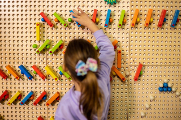 LiteZilla KidWorx™ Instant Reset Domino Dash Wall Activity, a young girl playing with colorful wooden dominoes on a pegboard wall.