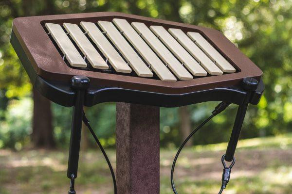 Freenotes Early Childhood Ensemble Musical Playground Set, a small outdoor musical instrument with a wooden keyboard mounted on a black metal stand, set in a park environment.