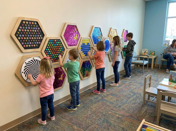 LiteZilla Multi-Sensory Hexagon Panel - Fidget Spinner XL, children playing with colorful sensory hexagon panels on a wall in a therapy or activity room.