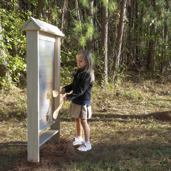 Wood Designs Junior Outdoor Thunderwall, a children’s outdoor educational and play wall with a clear panel and wooden frame, in a forest setting with a young girl engaging with it.