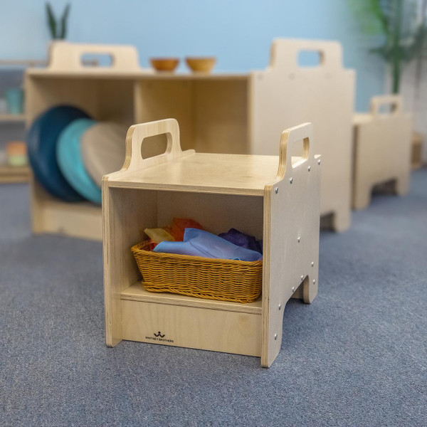 Whitney Brothers Stack-Able™ Seat, wooden modular children's seating with a basket of colorful fabrics inside, in a classroom setting.