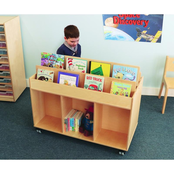 Whitney Brothers Mobile Book Storage Island, wooden children's bookshelf on wheels filled with children's books and a plush toy, in a classroom setting with a child looking at the books.