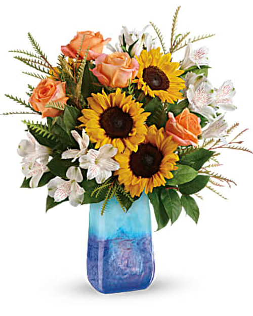This magnificent arrangement of golden sunflowers and orange roses, presented in a hand-blown art glass vase, brings the beauty of an ocean sunset to any occasion.   Light orange roses, white alstroemeria, and medium yellow sunflowers are arranged with grevillea and lemon leaf.
