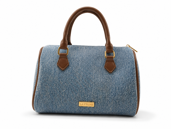 The Denim Boston Bag features soft, misty blue light wash denim, premium dark brown vegan leather trim, and polished gold-tone hardware.