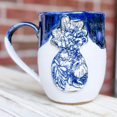 Exclusive Blue & White Pineapple Mug by Alabama Vitrified, handcrafted by Alabama artist Cari Cleckler. White pottery mug featuring a raised pineapple design finished in a classic blue-and-white motif, symbolizing hospitality, welcome, and Southern style. Available exclusively from Pine Apple Mercantile Company.