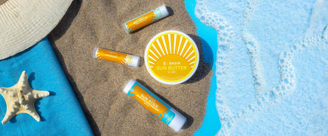 The image showcases Basin's sun care products playfully arranged on a sandy beach towel with a starfish, evoking a summery, seaside vibe. The products include their Sun Butter balm, SPF 30 Sun Stick lip balm, and SPF 30 mineral sunscreen.