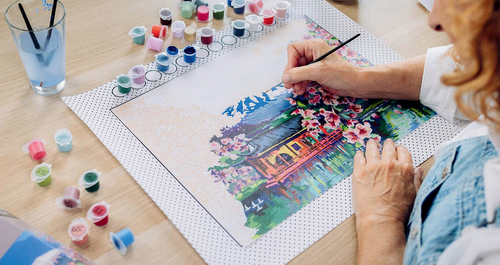 A pair of hands paints a CreArt paint-by-number Canvas Edition on the canvas fabric and protective surface mat