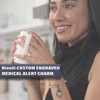 A smiling woman with dark hair holds a white mug with both hands, showcasing a silver medical alert charm bracelet on her right wrist. The bracelet features a heart-shaped charm with a gold medical alert symbol (star of life with a caduceus) in the center. She wears a dark top, and her light pink nail polish adds a subtle touch of style. The background is blurred, suggesting a modern indoor setting. A text overlay in the lower-left corner reads "Divoti CUSTOM ENGRAVED MEDICAL ALERT CHARM" in bold white font on a dark blue-gray background. The focus highlights the charm's design and functionality. A smiling woman with dark hair holds a white mug with both hands, showcasing a silver medical alert charm bracelet on her right wrist. The bracelet features a heart-shaped charm with a gold medical alert symbol (star of life with a caduceus) in the center. She wears a dark top, and her light pink nail polish adds a subtle touch of style. The background is blurred, suggesting a modern indoor setting. A text overlay in the lower-left corner reads "Divoti CUSTOM ENGRAVED MEDICAL ALERT CHARM" in bold white font on a dark blue-gray background. The focus highlights the charm's design and functionality.
