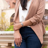 A close-up of a person wearing a decorative silver bracelet on their right wrist. The bracelet features an elongated oval panel with intricate filigree patterns and a polished central element, connected by links and a small dangling charm. The person is dressed in a light brown blazer, a white blouse, and dark blue jeans, with their right hand gently resting on their left. The background is softly blurred, showing hints of greenery, a stone or brick architectural element, and a light-colored ledge, creating a natural and elegant setting. A close-up of a person wearing a decorative silver bracelet on their right wrist. The bracelet features an elongated oval panel with intricate filigree patterns and a polished central element, connected by links and a small dangling charm. The person is dressed in a light brown blazer, a white blouse, and dark blue jeans, with their right hand gently resting on their left. The background is softly blurred, showing hints of greenery, a stone or brick architectural element, and a light-colored ledge, creating a natural and elegant setting.