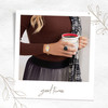 A lifestyle image framed like a Polaroid with decorative leaf motifs and the text "good time" at the bottom. The central focus is a person wearing a ribbed brown top, holding a white mug with black text partially visible, containing a dark liquid. The person wears a gold chain bracelet with a two-toned oval plaque featuring silver filigree and a gold sun-like design, and a square black gemstone ring on the right hand. A black-and-white patterned scarf drapes over their shoulder, and a pleated skirt with black, gray, and white vertical stripes is visible. The cozy aesthetic emphasizes warmth, style, and relaxation.