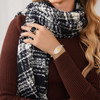 A close-up of a person wearing a dark brown ribbed sweater and a black-and-white patterned scarf with a fuzzy texture, draped around their neck and shoulders. The person’s right hand rests on their chest, showcasing a silver ring with a large, square black gemstone. On their right wrist, a silver and gold bracelet is visible, featuring a central oval design with intricate silver filigree and a gold starburst motif. Long, wavy blonde hair cascades over their shoulder, adding softness to the warm and stylish autumn or winter look. The lighting highlights the textures of the clothing and the shine of the jewelry.