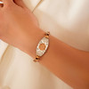 A close-up of a forearm wearing an elegant two-toned bracelet. The bracelet features a central oval silver-toned plaque with intricate floral or leaf patterns and a raised gold-toned hexagonal centerpiece displaying a four-leaf clover design. The chain consists of small, rounded, heart-shaped gold-toned links on either side of the plaque. The arm rests against a soft, creamy white fabric background with gentle folds, creating a warm and sophisticated presentation. A close-up of a forearm wearing an elegant two-toned bracelet. The bracelet features a central oval silver-toned plaque with intricate floral or leaf patterns and a raised gold-toned hexagonal centerpiece displaying a four-leaf clover design. The chain consists of small, rounded, heart-shaped gold-toned links on either side of the plaque. The arm rests against a soft, creamy white fabric background with gentle folds, creating a warm and sophisticated presentation.