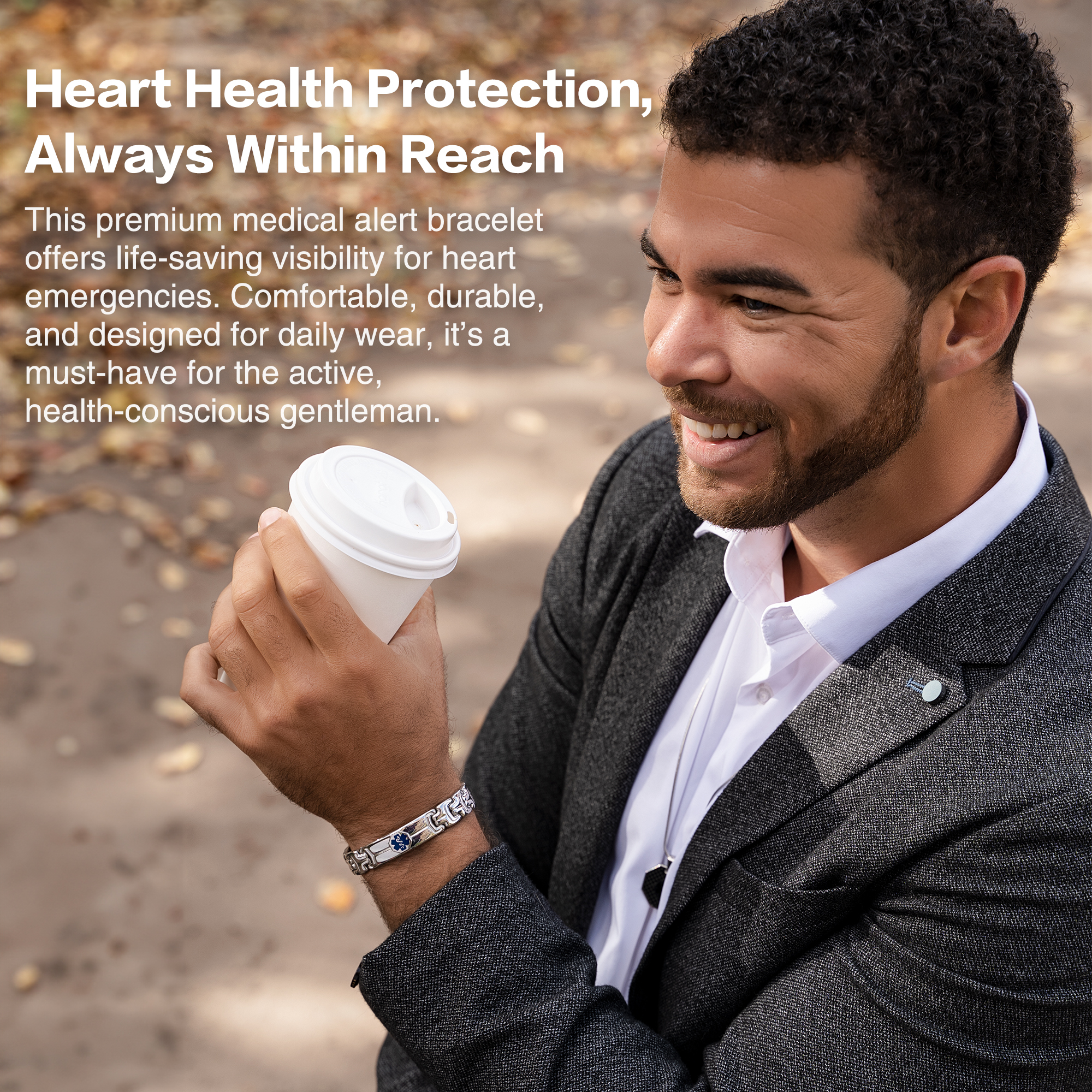 A smiling man with dark curly hair and a beard, wearing a white collared shirt under a dark grey blazer, holds a white coffee cup in his left hand. On his left wrist, he wears a silver medical alert bracelet with blue rectangular elements featuring a medical symbol. The background is blurred with warm autumn tones, and white text in the upper-left corner reads, "Heart Health Protection, Always Within Reach," promoting the bracelet's comfort, durability, and life-saving visibility.