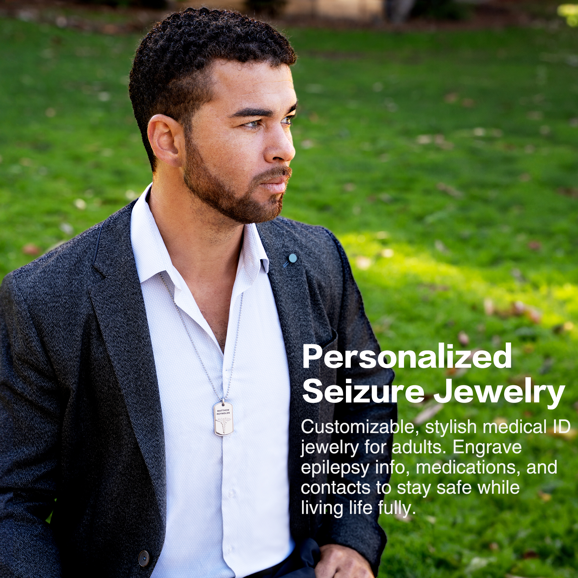 A stylish man with short curly dark hair, a beard, and light-colored eyes is wearing a white textured collared shirt and a dark gray blazer. Around his neck is a silver chain necklace with a rectangular pendant engraved with the name "Matthew Reynolds" and a caduceus medical symbol. The background is blurred green grass, suggesting an outdoor setting. On the right side of the image, white text reads, "Personalized Seizure Jewelry," followed by a description highlighting customizable, stylish medical ID jewelry for adults, designed to engrave epilepsy information, medications, and emergency contacts for safety and peace of mind.