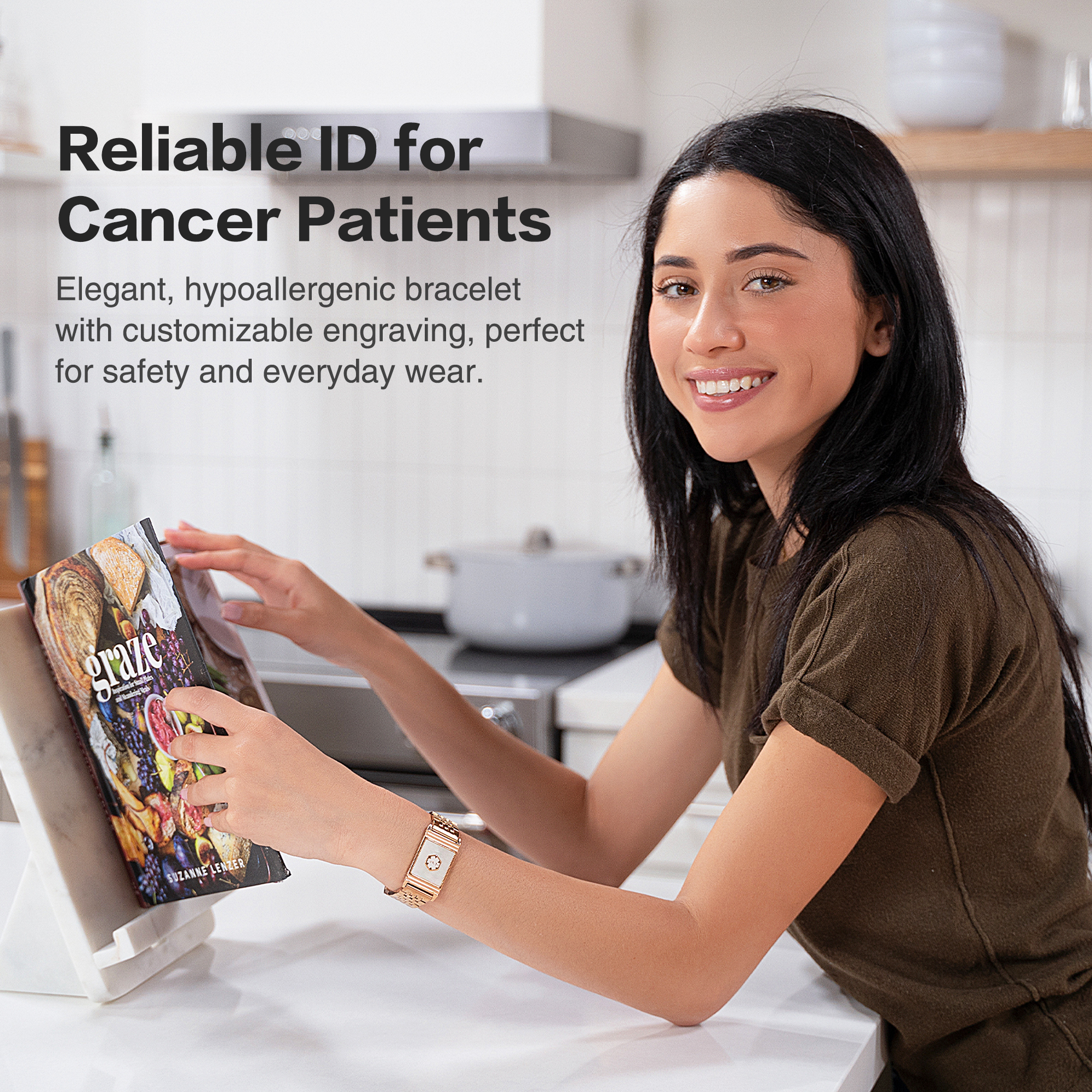 A smiling woman with dark hair wearing a gold-colored medical ID bracelet featuring a white center with a four-petal flower design. She is seated at a white kitchen counter, holding an open cookbook titled "graze" with colorful fruits and vegetables on the cover. The background shows a modern kitchen with a white range hood and a pot on the stove. Text overlay highlights the bracelet as an elegant, hypoallergenic, and customizable ID for cancer patients, perfect for safety and everyday wear.